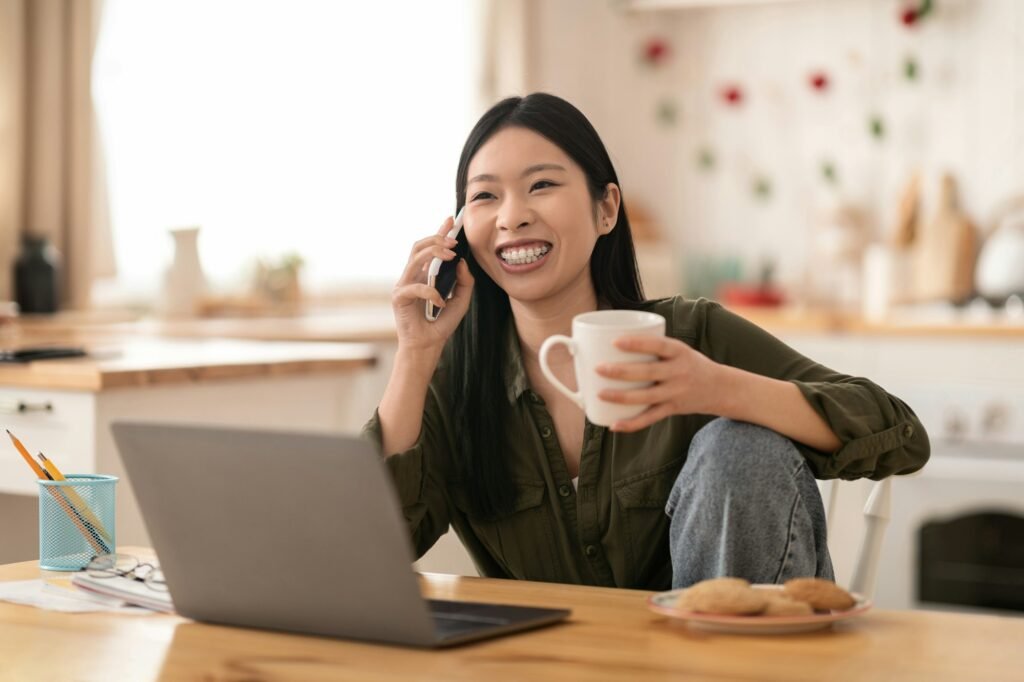 Cheerful korean woman using laptop at kitchen, talking on phone